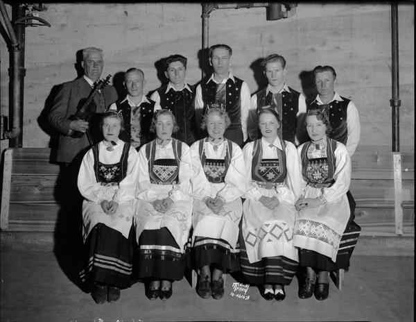 Group portrait of ten Norwegian folk dancers in costume, coached by John Odden, and Harold Smedal holding a Hardanger violin, who will perform for the Sons of Norway's Syttende Mai celebration.