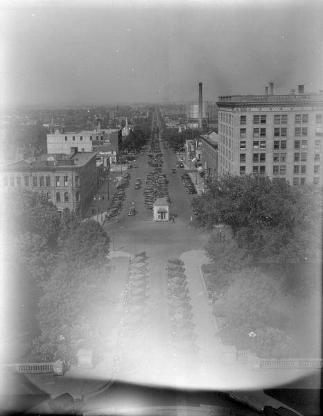 Elevated view of East Washington Avenue taken from an upper level of the Wisconsin State Capitol. Shows the First National Bank Building, American Exchange Bank, and other buildings. There is a smokestack further down the street on the right. Many automobiles are parked at an angle in the center of the street, as well as along the curbs.