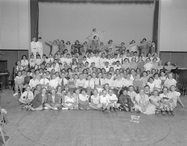 Group portrait of Franklin School students sitting and standing in front of and on a stage. There is a row of students on stage who are wearing togas and holding signs for the PTA program.