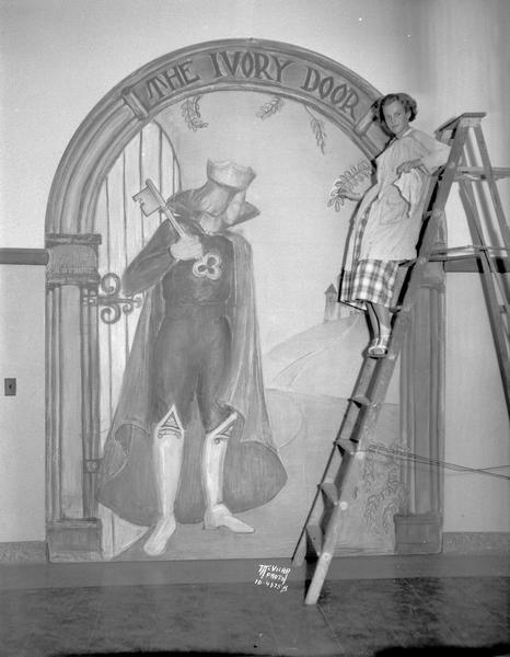 Jean Randolph, with bandaged ankle, which she sprained when she fell from the ladder on which she is standing, drawing an 8' x 12' crayon mural of a knight for the senior class play "The Ivory Door" at West High School.