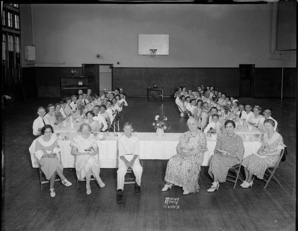 Five women and group of eighth grade students sitting around banquet tables at the Franklin School Mothers Club eighth grade banquet in the school gymnasium.