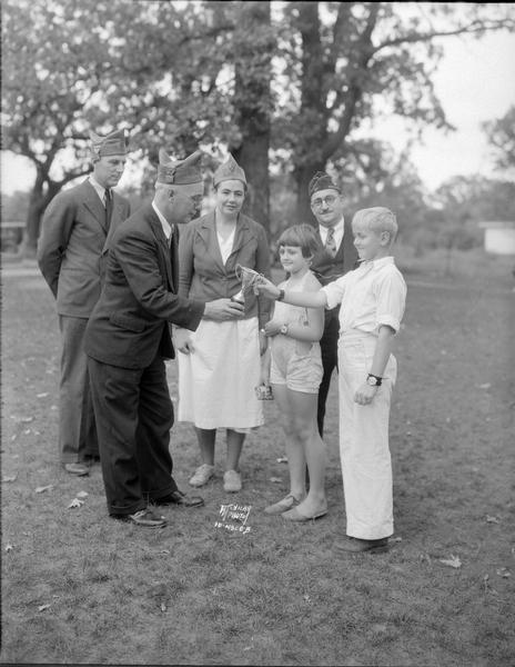 James L. Clarke, chef de gare of the American Legion 40 et 8 presents the silver cup trophy to Johnnie, Kiddie camper boy who gained the most weight at camp; he is also wearing his Boy Scout wristwatch prize. Between Mr. Clarke and Johnnie is Edith, Kiddie camper girl who gained the most weight, wearing her prize Mickey Mouse wristwatch. In the rear left to right are: Victor Ellis, Grand Cheminot of the 40 et 8; Ruth Coe, Kiddy camp director and Dr. M.J.J. Colluccy, commander-Elect fo the 40 et 8. Kiddie Camp located at 3910 Mineral Point Rd.