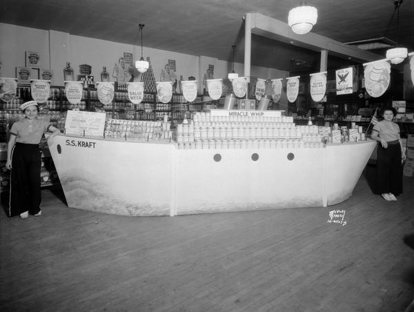 Two sales clerks dressed as sailors standing beside the ship S.S. Kraft filled with Kraft and Miracle Whip salad dressing products. The display is in Frank Bros. grocery store, 613 University Avenue. They are celebrating Kraft Salad Week in Madison. Shelves of canned goods are in the background.