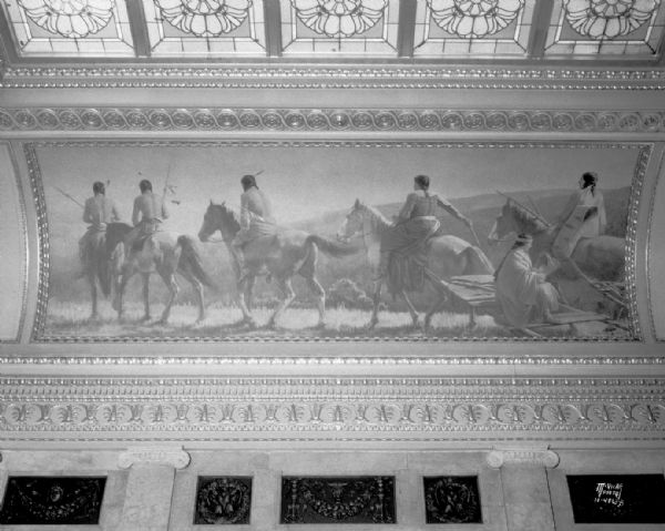 Mural depicting a party of Indians on horseback, on an upland, striking the trail (adapted from a photograph, by permission, made in the National Park). One of four murals by Charles Yardley Turner, representing the evolution of transportation in America. The stained glass skylight in the Wisconsin State Capitol North Hearing Room can also be seen. The title of the mural is: "Indians Striking the Trail."
