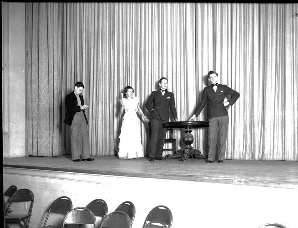 Four actors standing at a table on stage in the West High School auditorium.