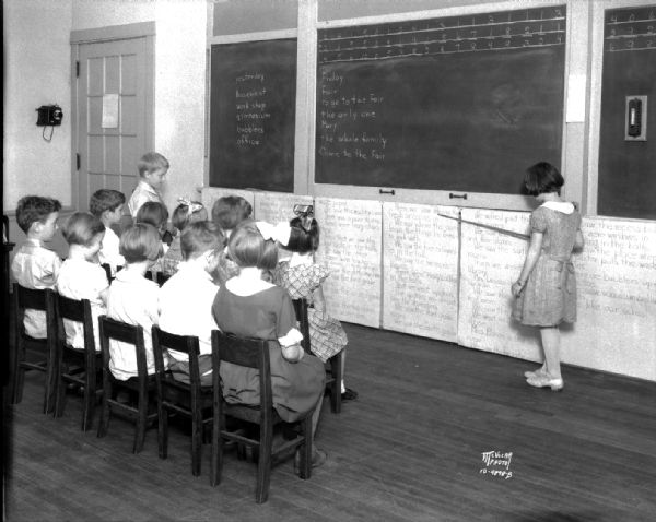 Randall School, 10 N. Spooner Street. There are children sitting in chairs reading from the blackboard, led by a student holding a pointer. Photograph taken for Miss Greenleaf, teacher.