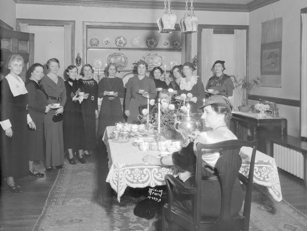 Group portrait of guests at the University League tea at the home of Dorothy Mendenhall, 205 N. Prospect Avenue. Sitting at the beautiful tea table and pouring were Ruth Page in foreground, and at the other end of the table Katherine R. Mead. In the background is a display of porcelain plates and platters.