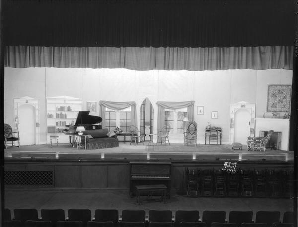 Stage of East High School set up with scenery for their play "The Importance of Being Earnest." Living room with grand piano, sofa, French windows, chairs, desk, fireplace. Below the stage is an upright piano and chairs stacked against the wall.