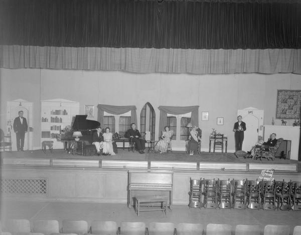 Cast of eight actors on the stage set with living room including piano, sofa, chairs, French windows, fireplace.