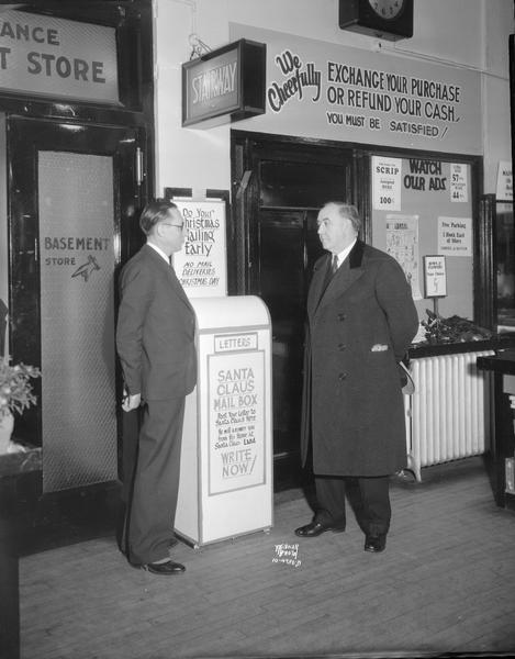 Harry Dimond, general manager of Hills Dry Goods Store & Postmaster Walter Hyland stand on either side of the "Santa Claus Mail Box" placed in Hill's Dry Goods store, 202 State Street at Christmas time. Sign says: "Post your letter to Santa Claus here. He will answer you from his home at Santa Claus Land. Write Now!"