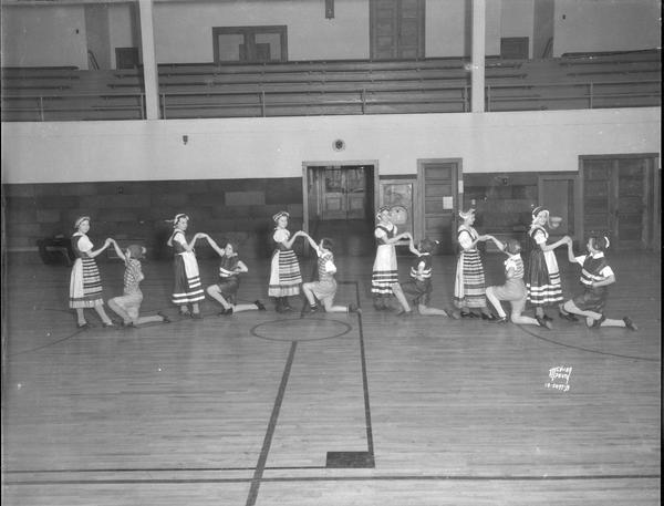 Nakoma School girls folk dance class. There are six "couples" in a row in ethnic costumes, with each "boy" on their knee holding a girl's hand.