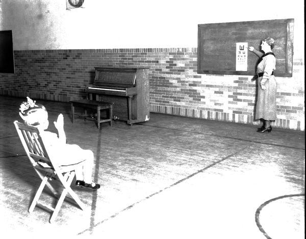 Woman pointing at chart and one child showing which way the E is pointing for Emerson School eye examinations. A piano is in the background.