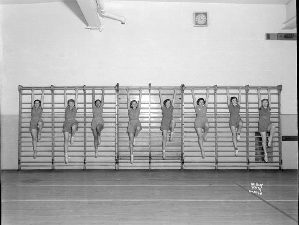 Eight Central High School girls in gym suits hanging by their arms from an exercise bar.