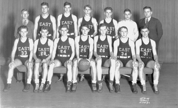 Group portrait of eleven male East High School basketball players in uniform, and a coach and two assistants.