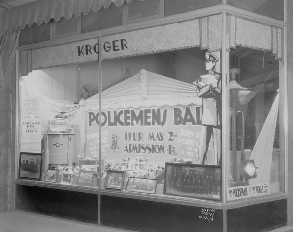 Kroger Grocery Store window, 3 North Pinckney Street, advertising policemen's ball. A stop and go light has been placed in the window with an officer (mannequin) brandishing a drawn gun standing guard. Includes photographs of the force from 1890 to the 1935 and a washing machine and groceries which will be given as prizes at the ball.
