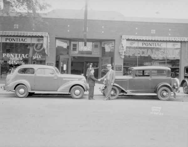 Waters Motors Co., 802 East Washington Avenue. Two men are shaking hands in front of old and new Pontiacs parked along the curb.