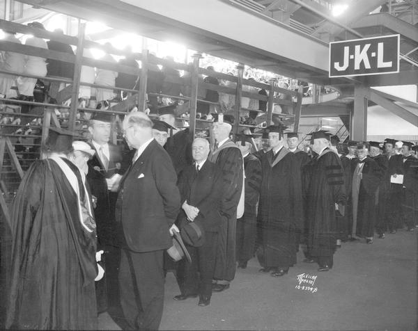 Lineup of professors and dignitaries in caps and gowns standing behind the bleachers preparing to march into the Field House for University of Wisconsin-Madison graduation ceremonies.