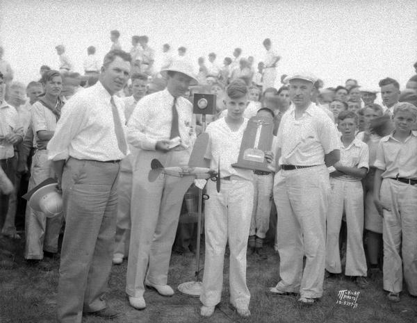Robert Grilley, holding his model airplane, receiving first place mahogany plaque in Junior Division of "Jimmy Allen" model airplane races at Royal Airport. He is surrounded by a crowd and three adult officials.