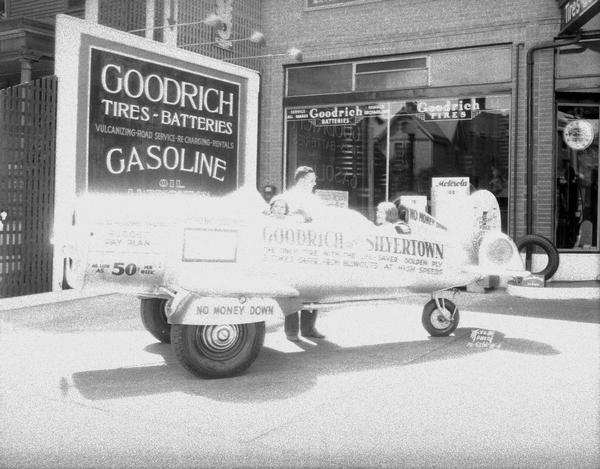 Man standing behind of two women sitting in a car that looks like an airplane parked in front of and advertising Goodrich Silvertown Stores, 515 University Avenue with the following sentence painted on the vehicle: "The only tire with the life-saver golden ply, three times safer from blowouts at high speeds."