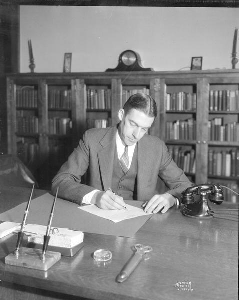 Rev. Edwin O. Kennedy sitting at a desk writing a letter, Christ Presbyterian Church, 124 Wisconsin Avenue. There is a telephone on the desk, and books are in a bookcase in the background.