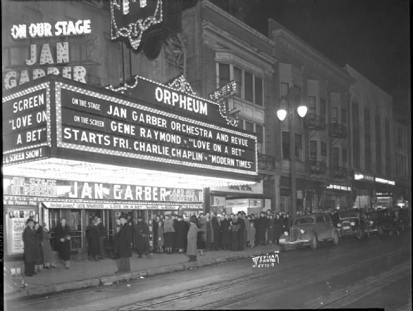 View from street towards a crowd lined up in front of Orpheum Theater to see Jan Garber orchestra and revue and Gene Raymond in "Love on a Bet,"  200 block of State Street showing many of the businesses.