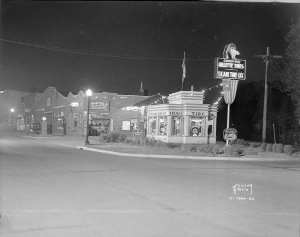 Night view of the Licari Tire Company station 767 West Washington Avenue, at the corner of West Main Street and West Washington Avenue. The Art Deco Trachte building has two American flags, and a neon sign has a bear stepping through a tire and "A Bear for Wear, Gillette Tires." Also shows Kennedy Dairy garage, 765 West Washington, and Zach Trotter's restaurant, 763 West Washington, with the Wisconsin State Capitol dome in background.