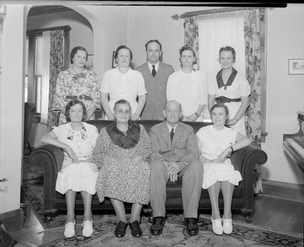 Group portrait of five adults standing behind four adults who are sitting on a sofa in a living room. All of the adults are members of the Dieder family. A piano is on the right.