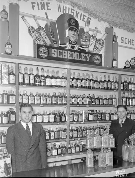 Two men are standing behind the counter in front of a wall display of Schenley Liquors on shelves at the Capitol Liquor Store, 217 State Street.