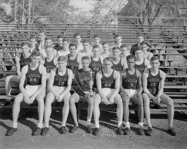 Outdoor group portrait of Central High School boys track team in uniform, with one member in the center holding a trophy.