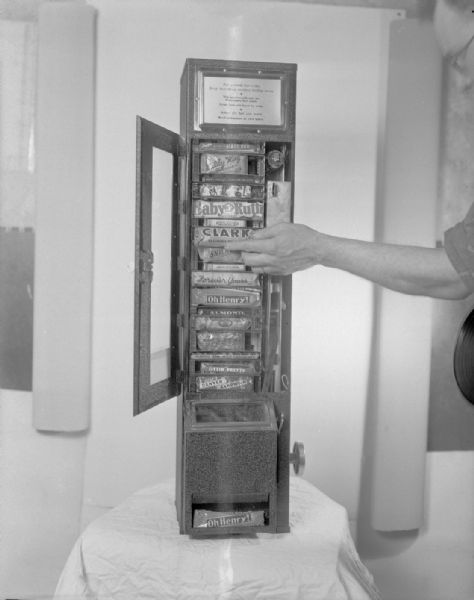 View of a man's hand reaching in to fill Coan Sletteland Co. 5 cent candy bars into a vending machine filled with name brand candy bars.