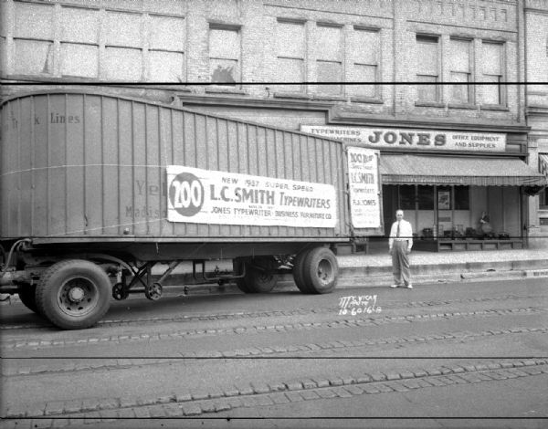 View towards one man standing beside a semi-trailer truck delivering 200 new 1937 super speed L.C. Smith typewriters to Jones Typewriter — Business Furniture Co., 506 State Street.