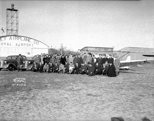 Outdoor group portrait of Legionnaires at the Royal Airport. Also shows two automobiles, an airplane and the Morey Airplane hanger.