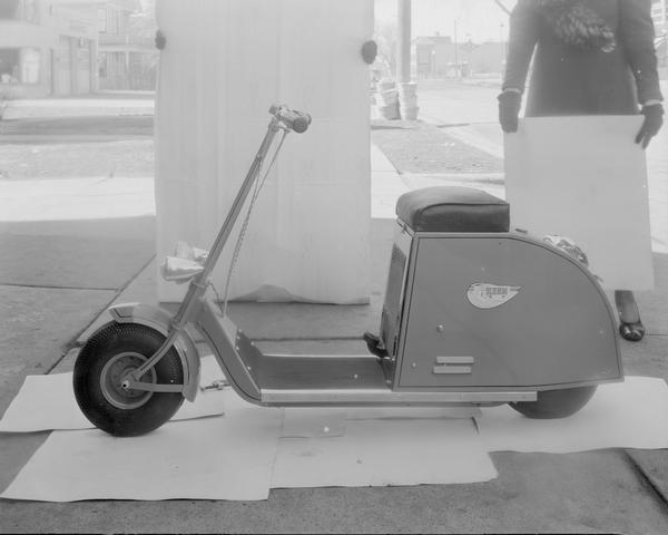 Keen Power Cycle, a streamline style motor scooter, made by Keen Manufacturing Co., 2117 Atwood Avenue, displayed outside on Atwood Avenue with two people holding up backdrops.