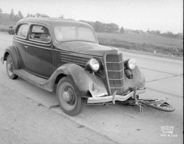 Accident scene on Highway 12-18 one-half mile east of the Dane County fairgrounds. Close-up of front of automobile, with bicycle under the car. Katherine Beglinger who was riding the bicycle was killed.