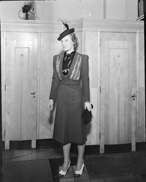 Model wearing hat, dress with jacket, spectator shoes, carrying purse and gloves, part of fashion series from Kessenich's Ready to Wear, 201-203 State Street, taken in front of dressing rooms.