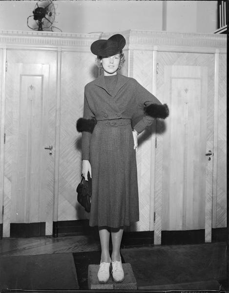 Model wearing hat, dress with jacket trimmed in fur, and carrying a purse, is standing on blocks in front of the dressing rooms at Kessenich's Dry Goods store, 201-203 State Street.