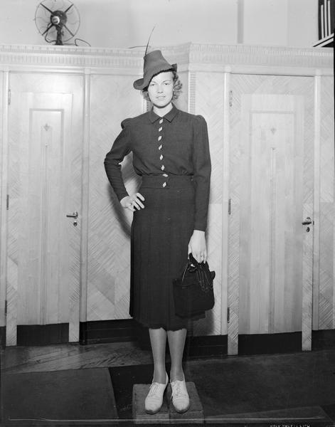 Model wearing hat, dress with jacket, carrying purse and gloves, and standing on blocks. Part of fashion series from Kessenich's Ready to Wear, 201-203 State Street, in front of dressing rooms.