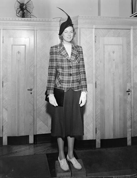 Model wearing hat, plaid jacket with plain skirt, wearing gloves, carrying purse, standing on blocks. Part of fashion series from Kessenich's Ready to Wear, 201-203 State Street, in front of dressing rooms.