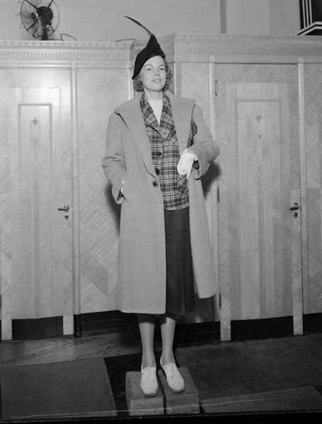 Model wearing hat and gloves, with coat over plaid jacket and plain skirt. The female model is standing on blocks. Part of fashion series from Kessenich's Ready to Wear, 201-203 State Street, in front of dressing rooms.