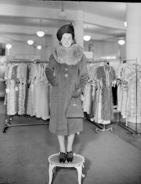 Model standing on step stool wearing hat and gloves, with fur collar coat, dress shoes, and carrying a purse. Part of a fashion series from Kessenich's Ready to Wear, 201-203 State Street, showing racks of dresses.