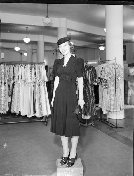 Model standing on box wearing hat and carrying gloves and purse, and wearing a dress. Part of a fashion series from Kessenich's Ready to Wear, 201-203 State Street, showing racks of dresses.