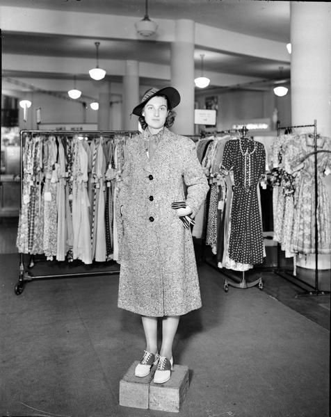 Model standing on box wearing hat and gloves, with tweed coat. Part of a fashion series from Kessenich's Ready to Wear, 201-203 State Street, showing racks of dresses.