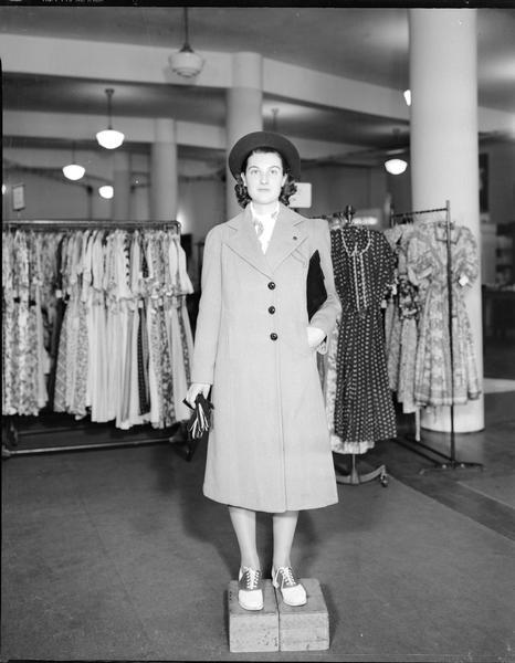 Model standing on box wearing hat and coat, carrying gloves and purse. Part of a fashion series from Kessenich's Ready to Wear, 201-203 State Street, showing racks of dresses.