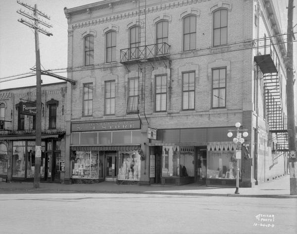 Fort Atkinson street corner showing, left to r.ight, a drug store, photography studio, C.B. Hinterschied 5 cent-$1.00 store, The Green Hat Lunches and Hopkins Shop for Men. Also shows lamppost and highway signs for Highways 12, 26 and 89.