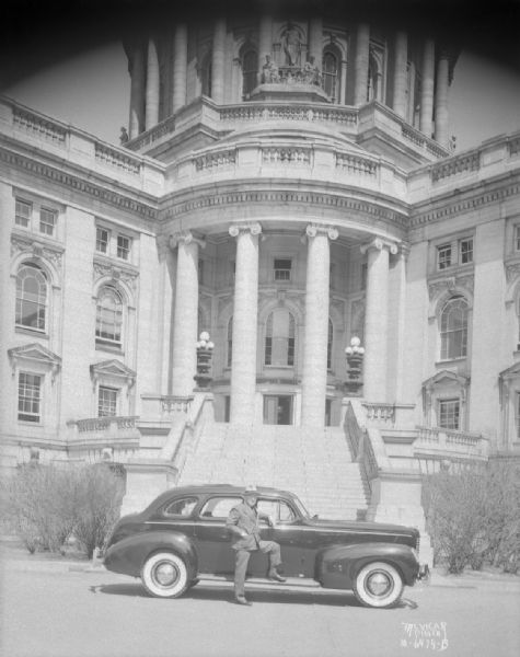 Governor Julius P. Heil is standing with his foot on the running board of a 1939 Nash Lafayette automobile, which is parked in front of the steps of the Wisconsin State Capitol.