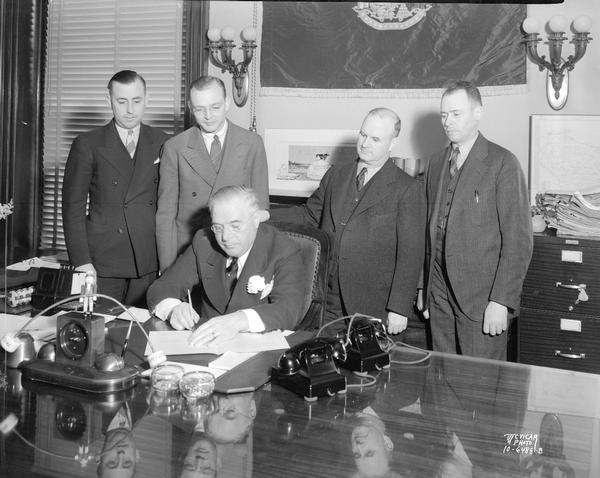 Governor Julius P. Heil sitting at his desk signing a bill, with four men standing and looking on.
