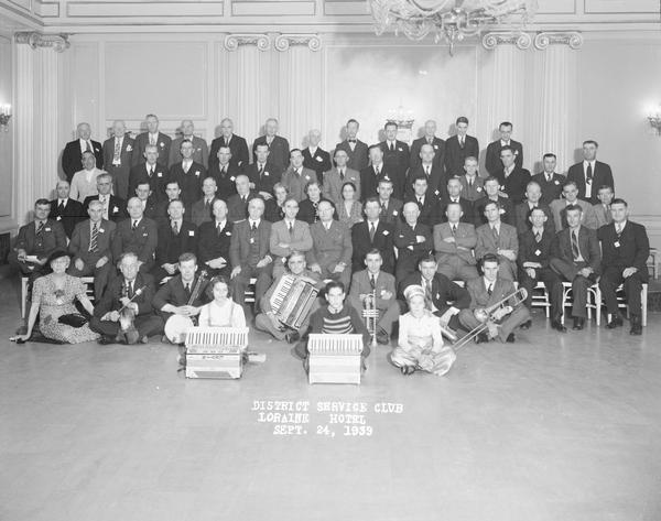 Group portrait of Milwaukee Railroad Service Club convention in Crystal Ballroom of Loraine Hotel. A group people are in front posing with musical instruments, including accordions, trombone, cornet, and banjo.