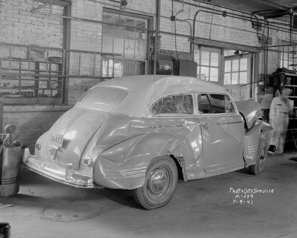 Three-quarter rear view from right of a damaged car in Schultz Body Co. garage, 1336 Regent Street. A mechanic is standing in the background.