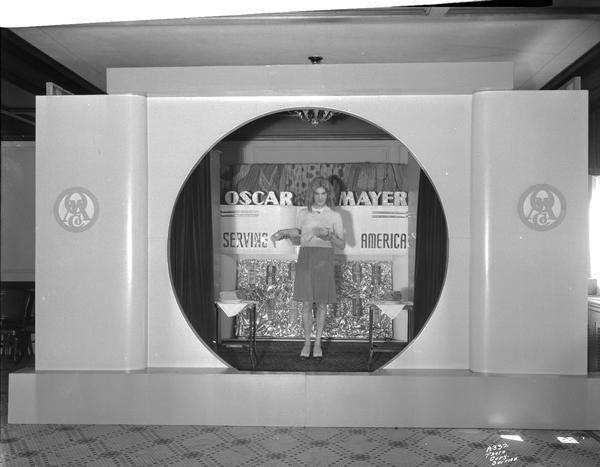 A female model is wearing short dress and is standing and holding an Oscar Mayer meat product as part of a meat products display.