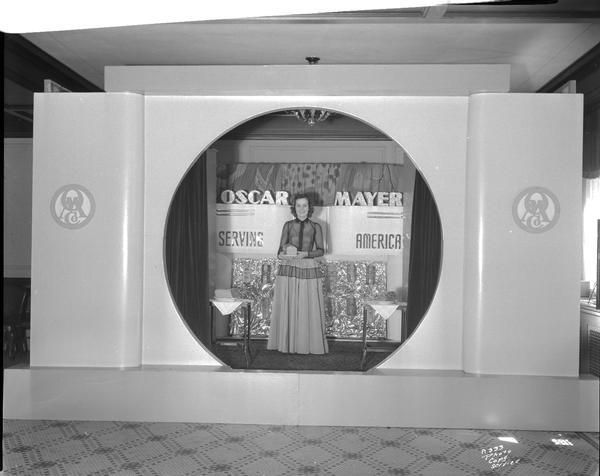 A female model is wearing a long dress and is standing in the "Oscar Mayer Serving America" stage set holding a meat product.
